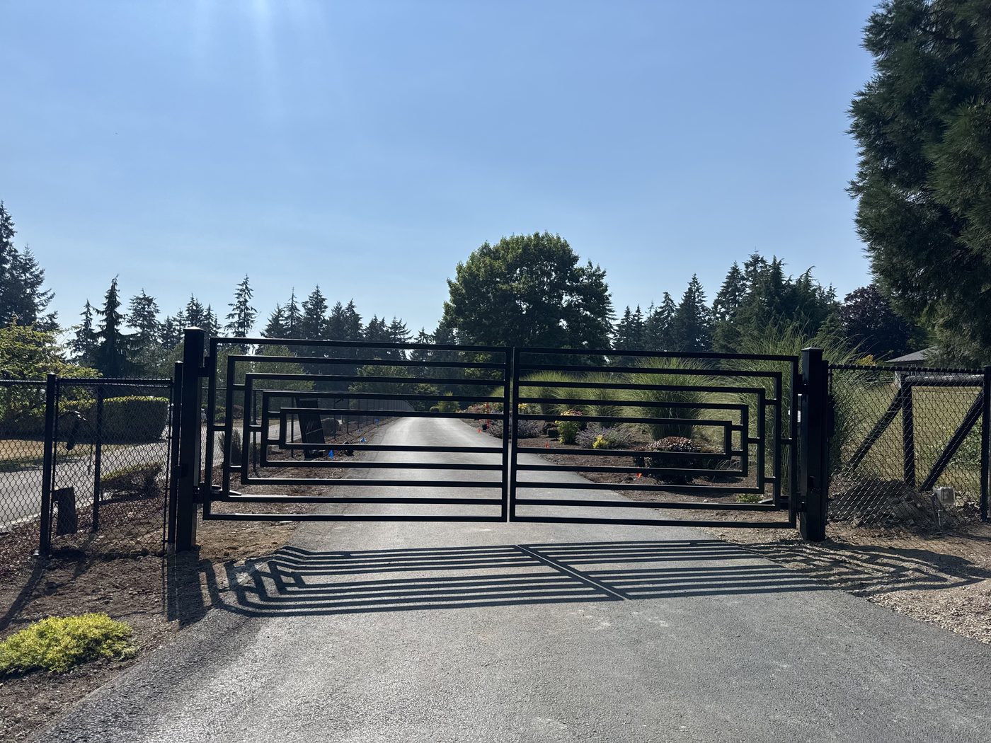 Black steel double driveway gate with a concentric rectangle design, closed across a gravel drive on a sunny day